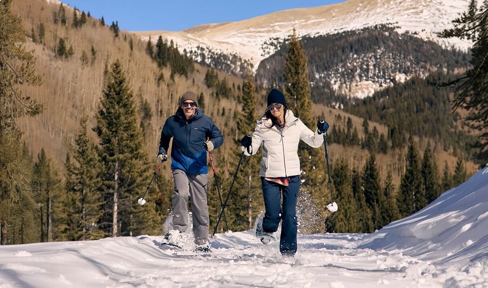 Two people snowshoeing along a snowy trail with mountain backdrop.